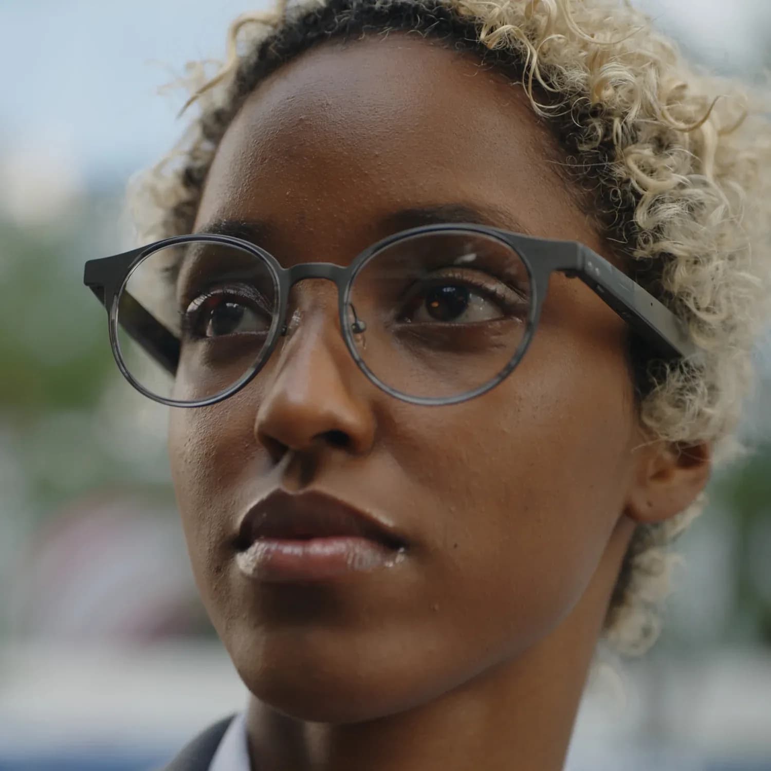 Dymesty AI Glasses en metal gris oscuro, montura redonda usada por una mujer de cabello rizado rubio al aire libre.