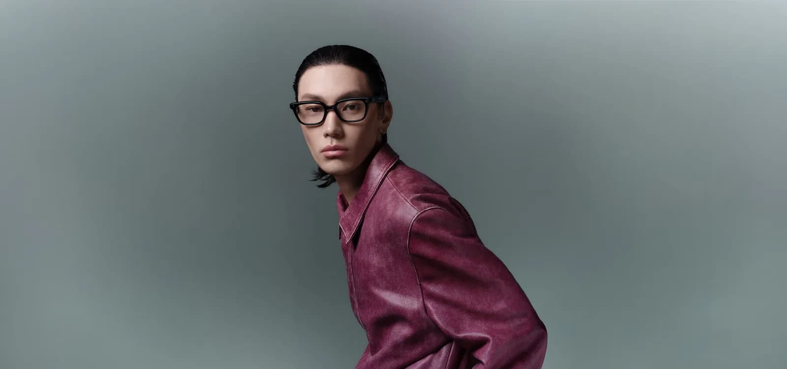 Gafas Gentle Monster Intelligent Eyewear en negro, montura cuadrada con cristales transparentes, puesta por un hombre con chaqueta de cuero granate, fondo gris.