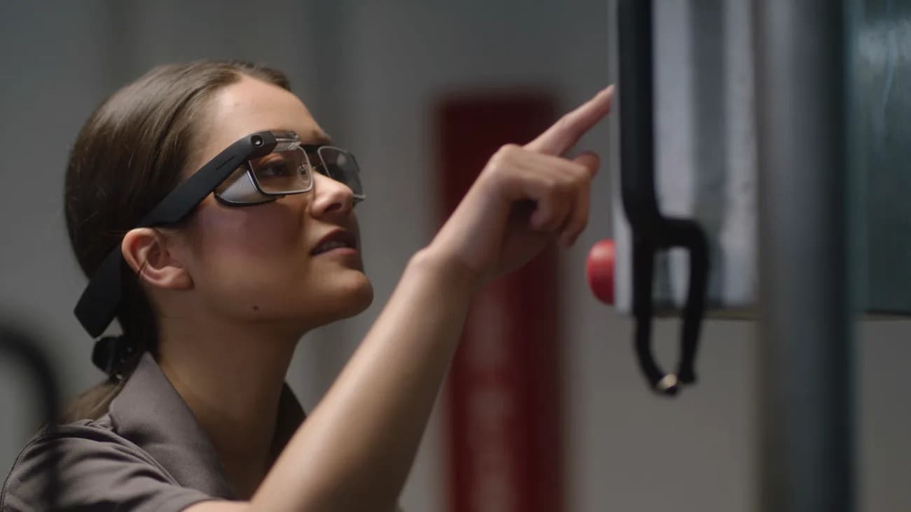 Gafas Google Glass Enterprise Edition 2 usadas por una mujer que señala una máquina, con la cámara y la pequeña pantalla visibles sobre su ceja derecha.