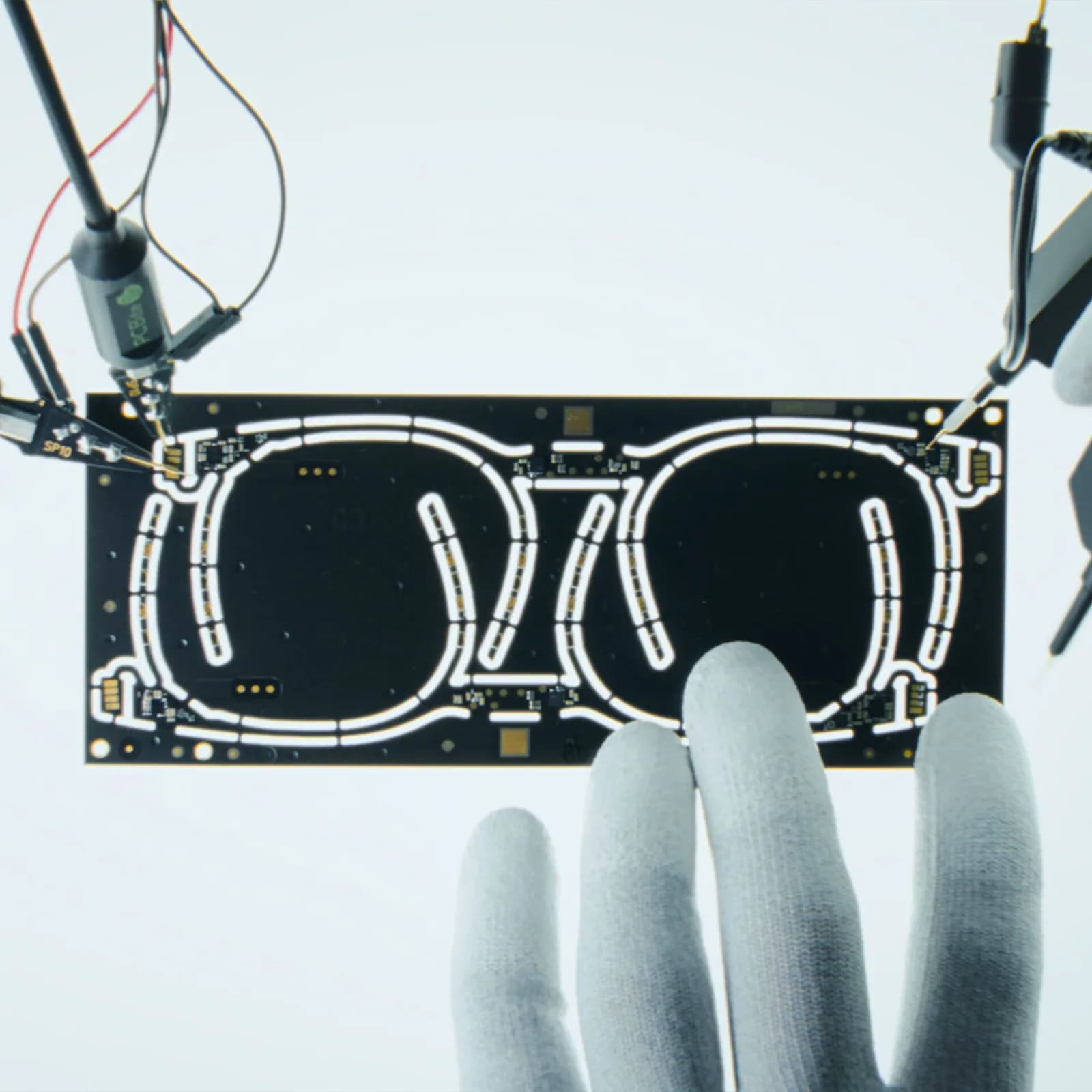 Placa de circuito del prototipo de las IXI Adaptive Eyewear con forma de gafas, conectada a sondas de prueba, sostenida por una mano con guante.