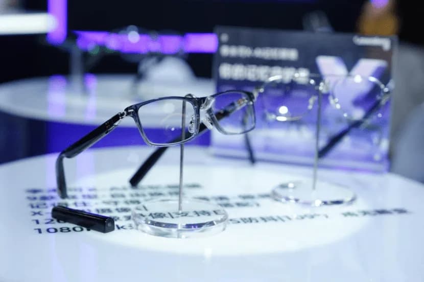 LAWK X-AI Memory Glasses in black, resting on a clear display stand at a trade show booth, with a rectangular half-rim frame.