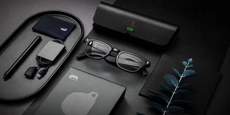Gafas B by Lenskart plegadas sobre una superficie oscura, junto a su estuche, caja, paño de limpieza y cable de carga.