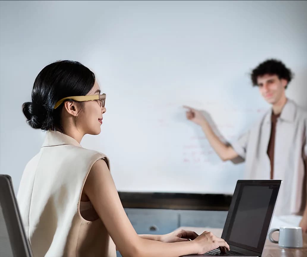 Gafas Meizu StarV Air2 en beige, en el rostro de una mujer escribiendo en un portátil, con un hombre señalando la pizarra al fondo.