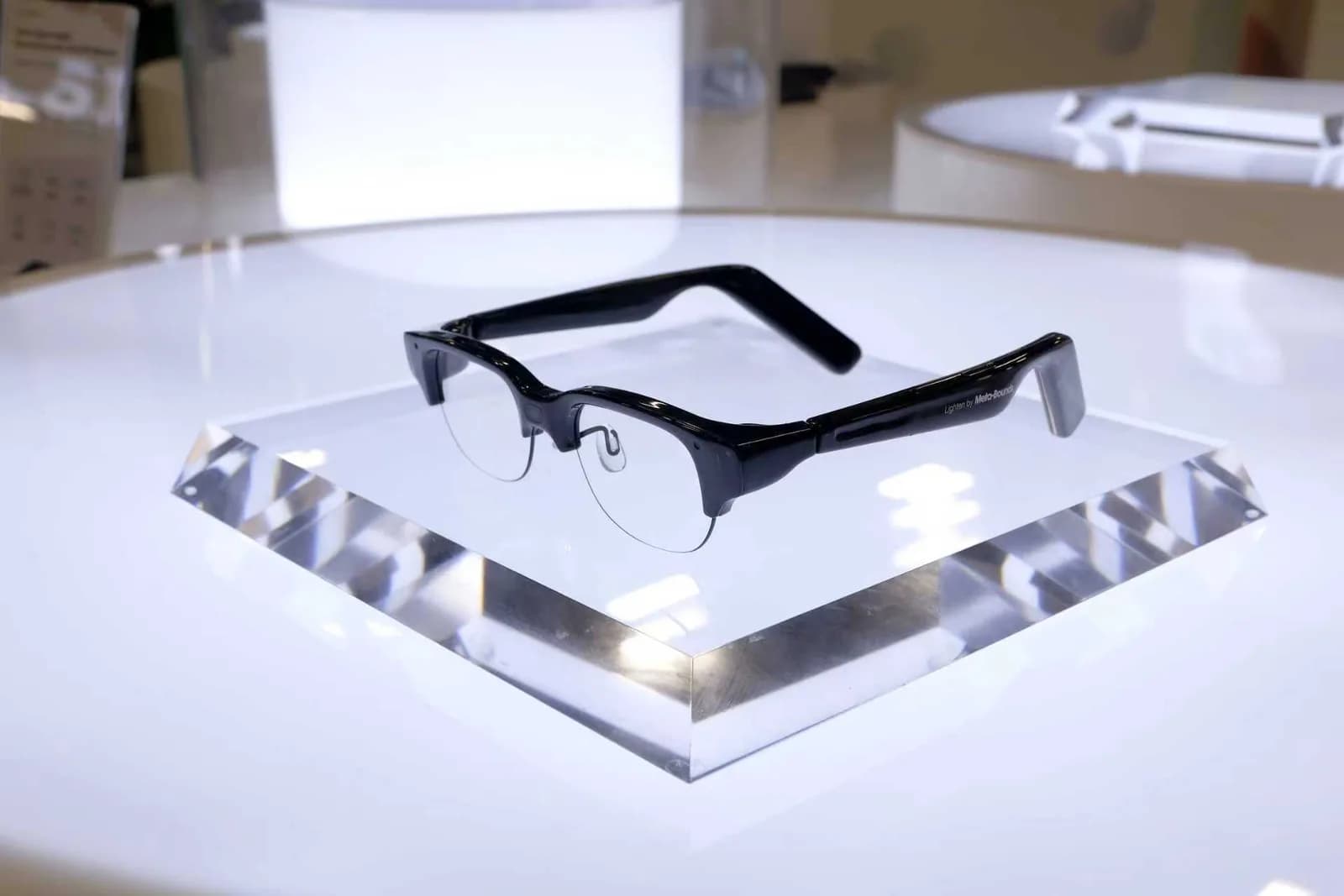Gafas Meta-Bounds AI+AR Glasses en negro, montura media al aire con aro inferior de alambre metálico, sobre un soporte de acrílico transparente en un stand.
