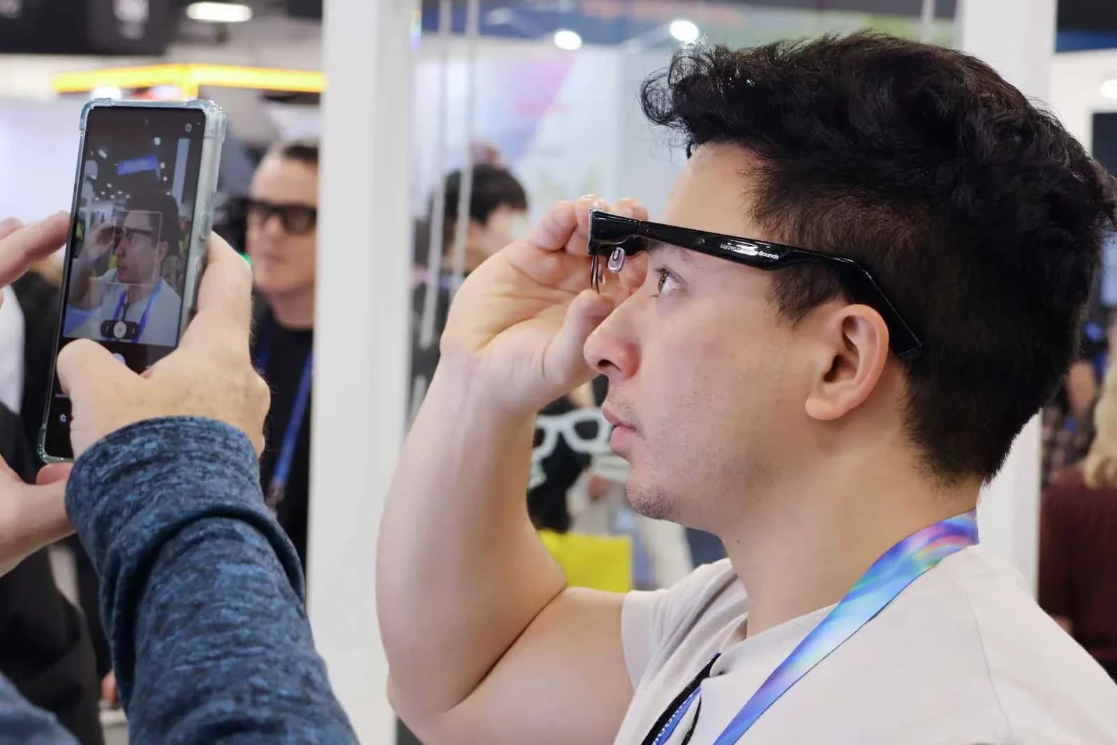 Óculos Meta-Bounds AI+AR Glasses em preto, armação meio-aro, sendo colocados no rosto de um homem numa feira, enquanto outra pessoa o fotografa com o celular.