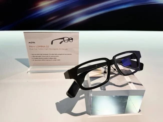 Gafas MOVA Yaomu G2 en negro, montura rectangular gruesa, apoyadas de pie sobre un bloque de vidrio transparente en una mesa blanca de exhibición.