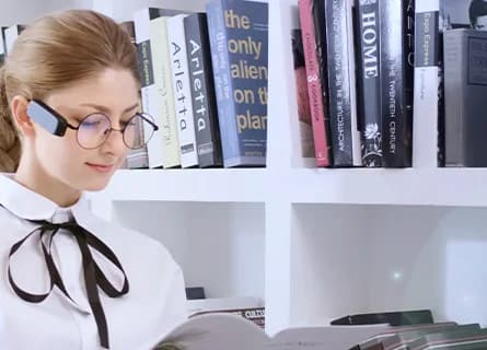 NextVPU AngelEye Smart Reader clipped to round glasses worn by a woman reading a magazine in front of a bookshelf.
