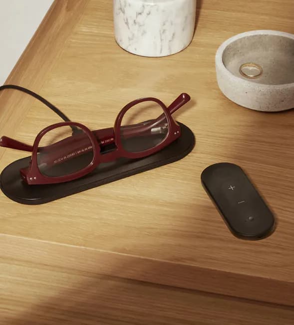 Nuance Audio Hearing Glasses in burgundy resting on a charging pad next to a black remote control on a wooden nightstand.