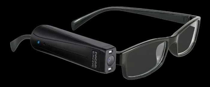 Módulo de cámara OrCam MyEye sujeto a la patilla izquierda de unas gafas rectangulares negras, con el objetivo y una luz azul indicadora visibles.