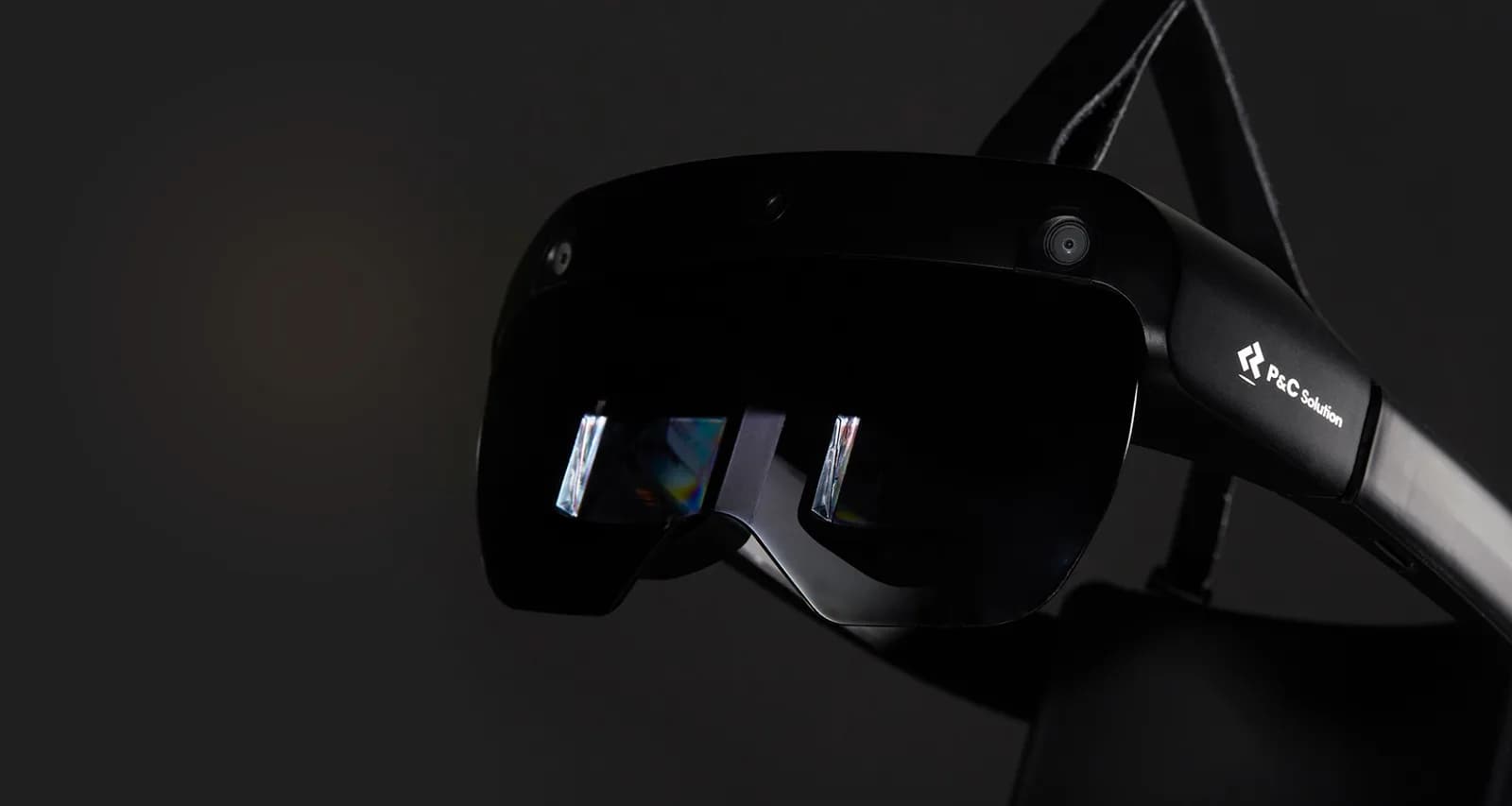 El METALENSE 2 visto de cerca en ángulo sobre fondo oscuro: un visor binocular negro con pantalla ahumada, dos cámaras en la cara frontal y una tercera entre ellas, los dos prismas de forma libre brillando dentro de la pantalla y un brazo rígido con la marca P and C Solution que llega hasta la cinta de cabeza.