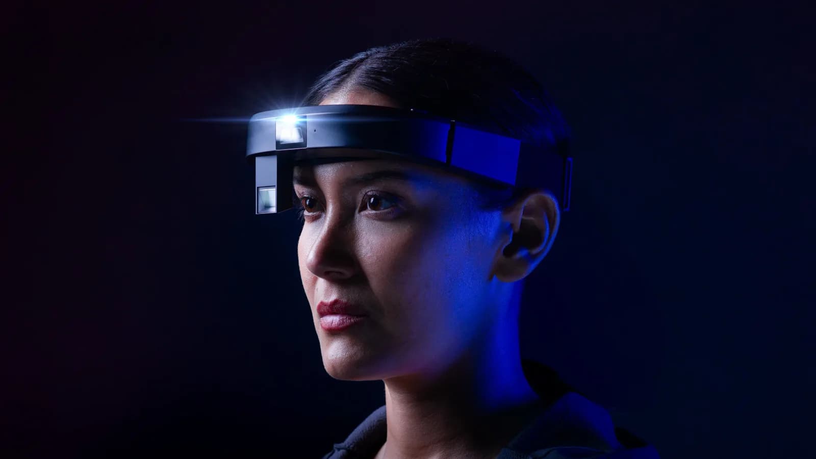 RealWear Arc 3 headset in black, worn by a woman in profile with the display arm lowered and glowing in front of her right eye.