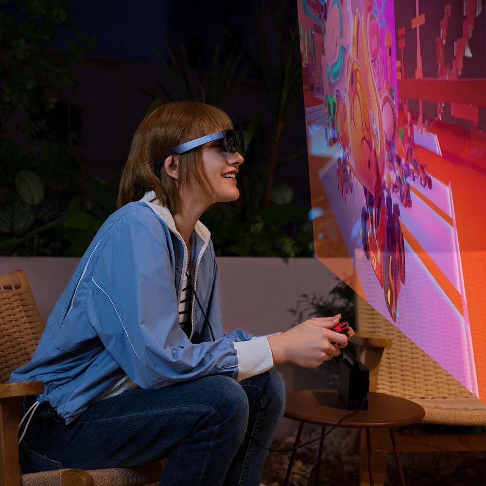 A woman wears black TCL NXTWEAR S+ glasses in profile, seated outdoors at night, holding a game controller.