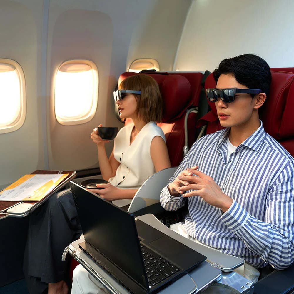 A man and a woman wear black TCL NXTWEAR S+ glasses side by side on an airplane, the man's pair connected to a laptop by a cable.