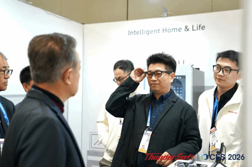 A man adjusts black-framed ThunderSoft TurboX AI Glasses on his face at a trade show booth, with colleagues standing nearby.