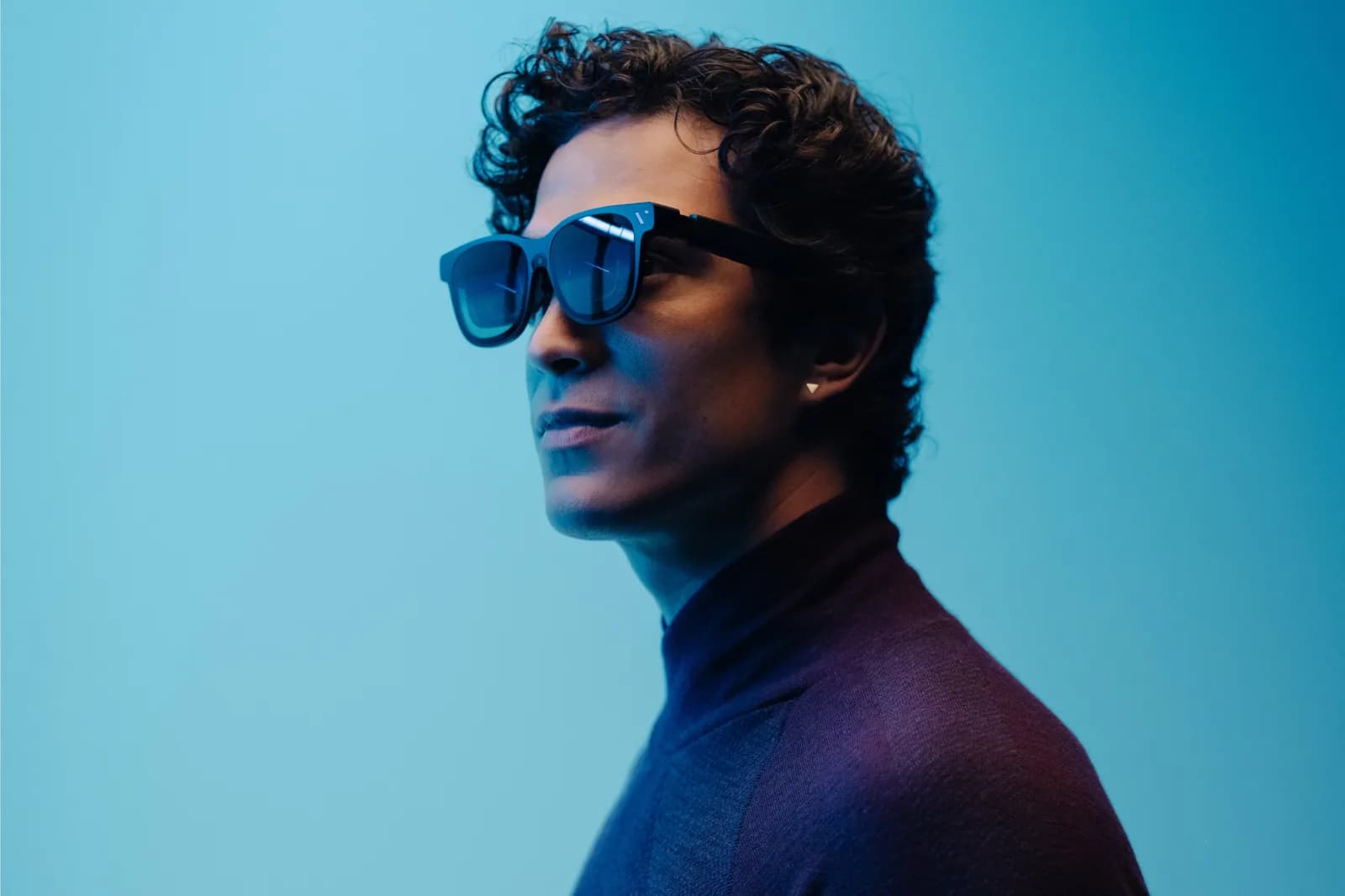 A man seen in profile against a teal and purple background wearing the Viture Beast: from the side the device reads as a pair of thick dark sunglasses, with the temple wider than an ordinary frame and a cable-free tip behind the ear.