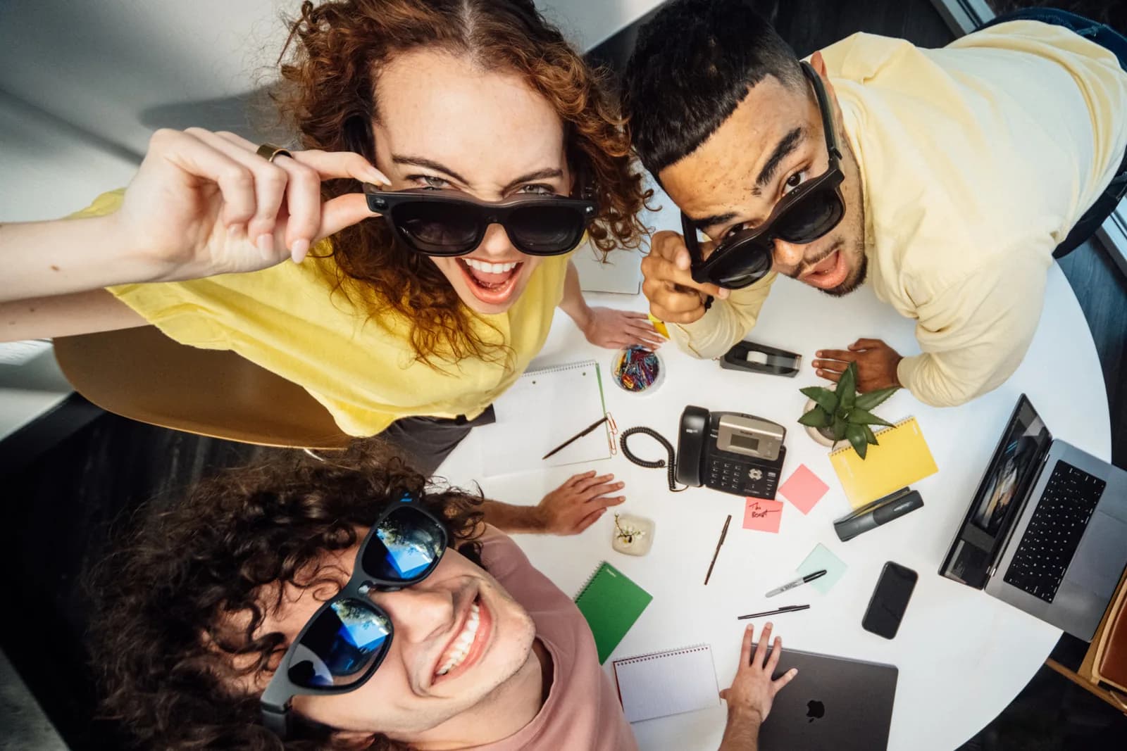 Three people leaning over a desk covered with notebooks, phones and sticky notes, all three wearing the Viture Beast and looking up at the camera; one of them is pulling the frame down the nose.