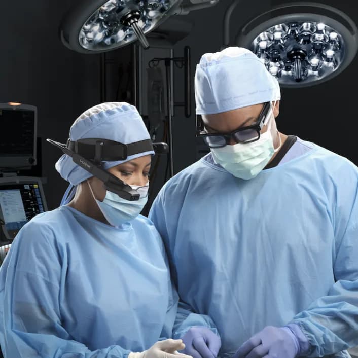 Dos personas con bata, gorro y mascarilla quirúrgicos trabajan bajo las luces de un quirófano; la de la izquierda lleva el Vuzix M4000 en una cinta de cabeza.
