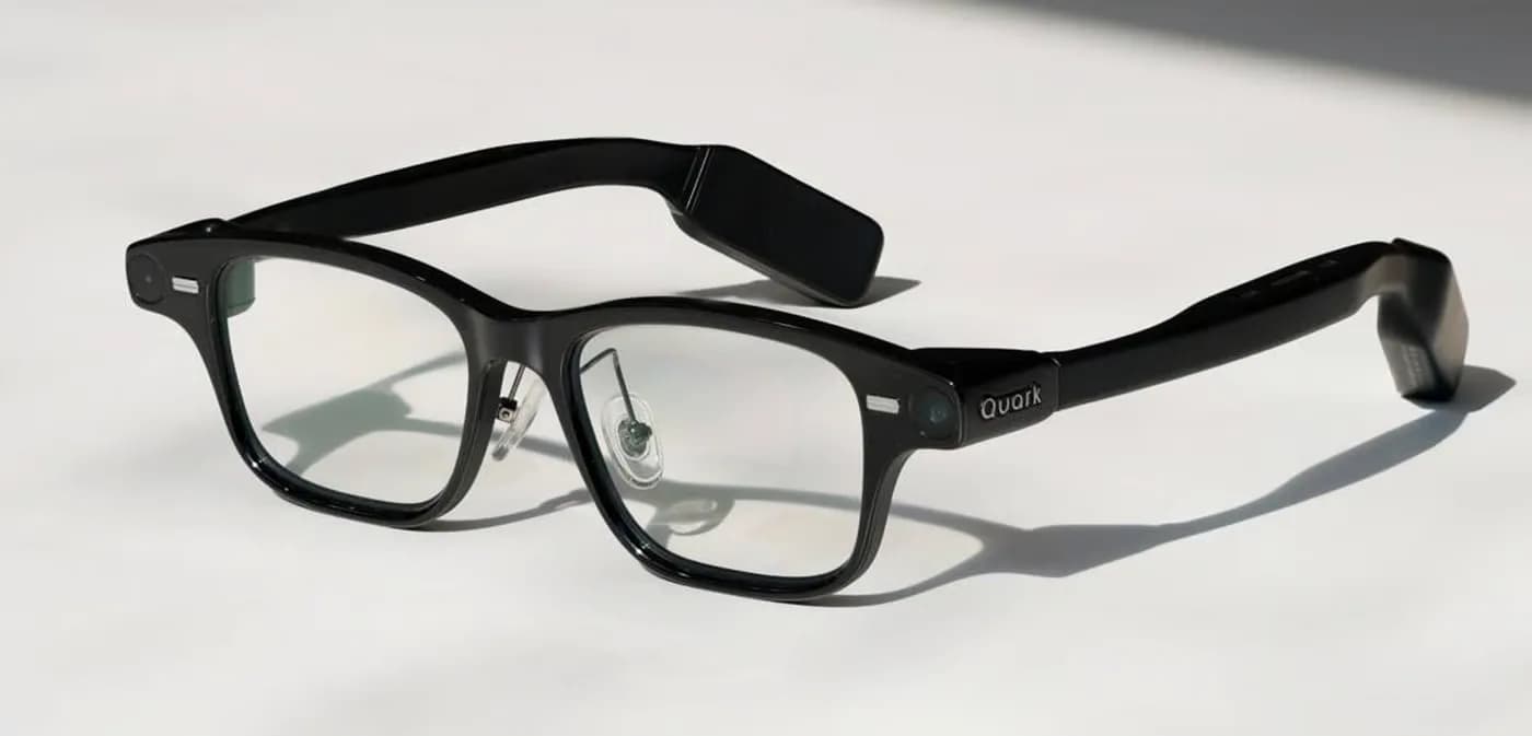 Quark AI Glasses S1 in black, resting on a white surface, with a thick rectangular frame, the Quark logo and a small camera lens.