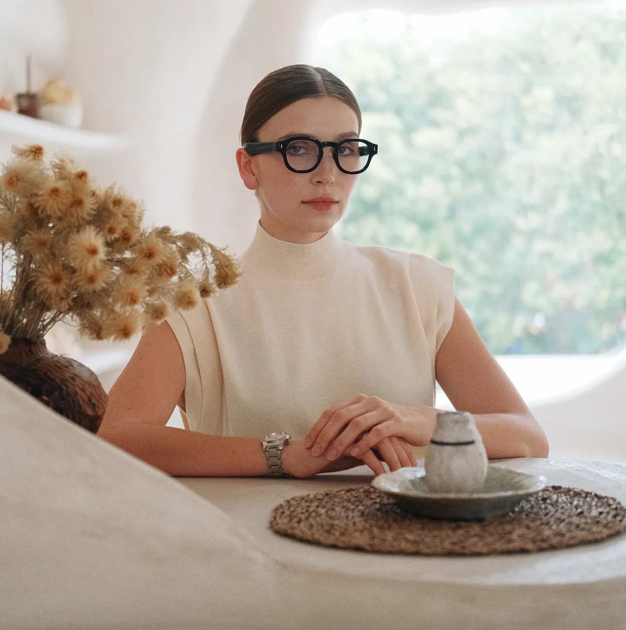 Halliday G2 smart glasses in black, round frame worn by a woman seated at a table near a window, dried flowers beside her.