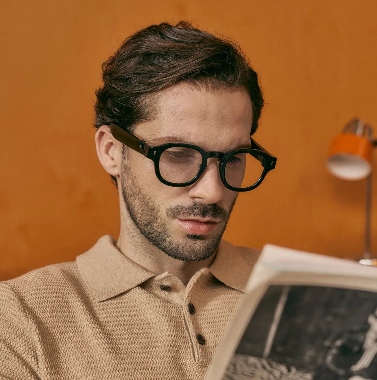 Halliday G2 smart glasses in black, worn by a man reading a book, round frame with clear lenses, warm orange background.