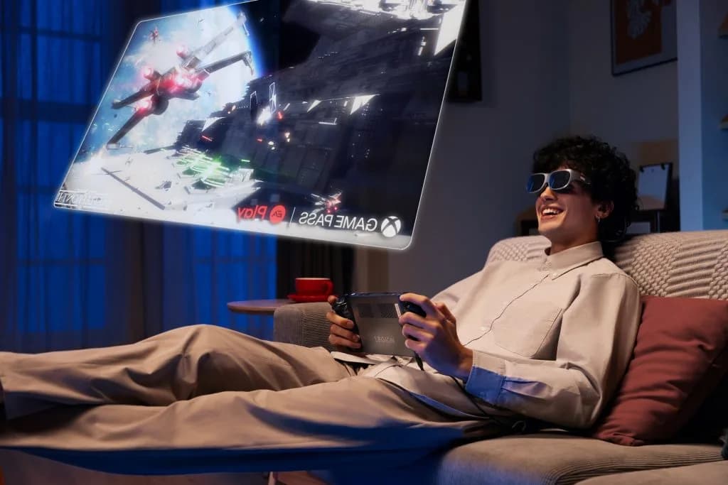 Lenovo Legion Glasses in black, worn by a person reclining on a sofa while holding a Legion handheld game console.