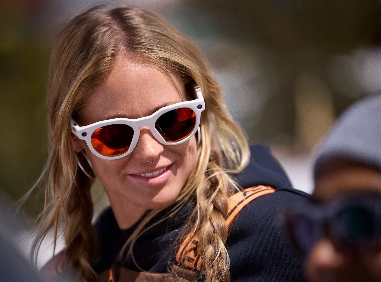 Oakley Meta HSTN glasses in white, round frame with orange mirrored lenses, worn outdoors by a smiling woman with braided blonde hair.