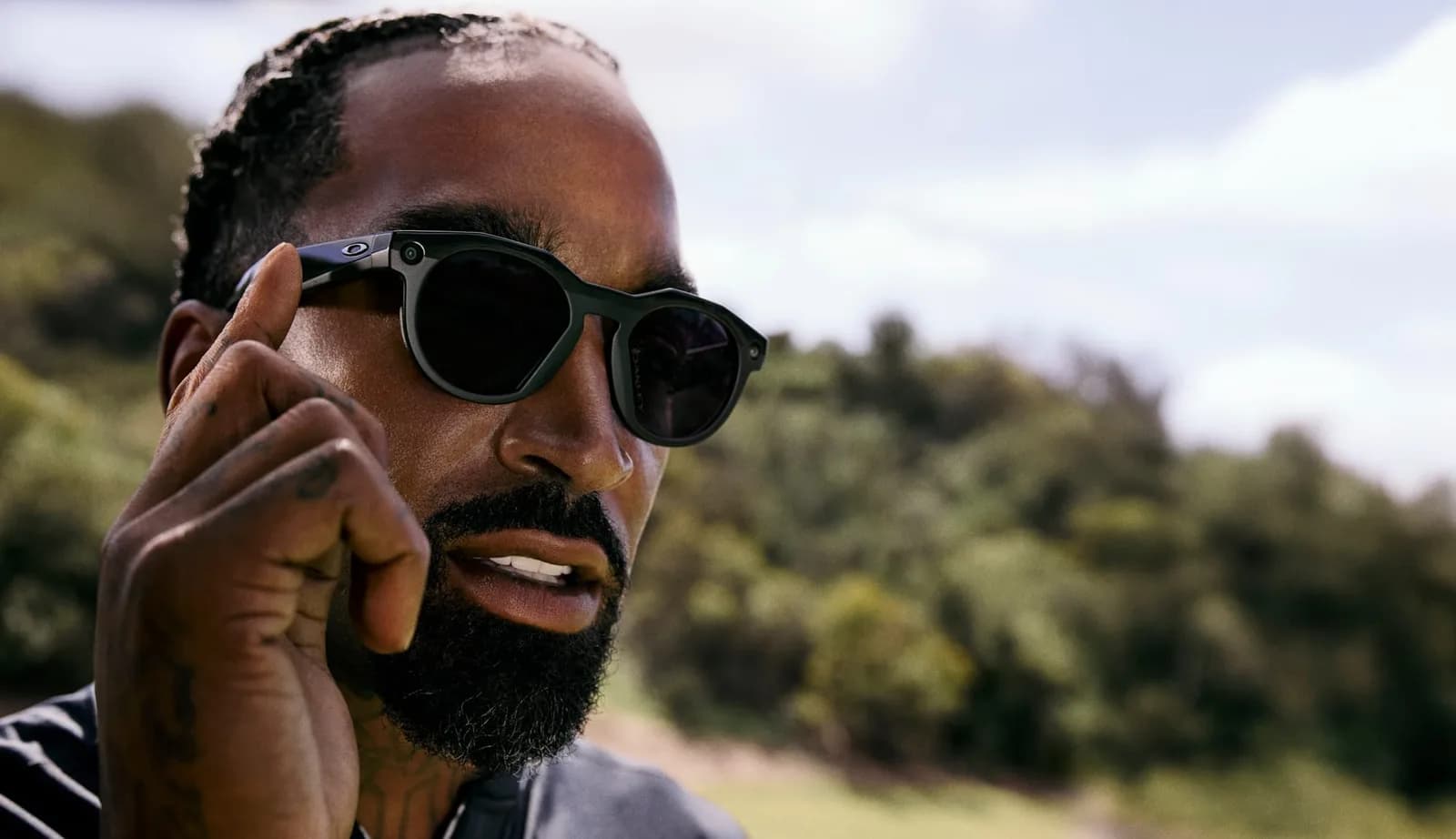 Oakley Meta HSTN glasses in black, round frame with dark lenses, worn outdoors by a bearded man adjusting them with his fingers.