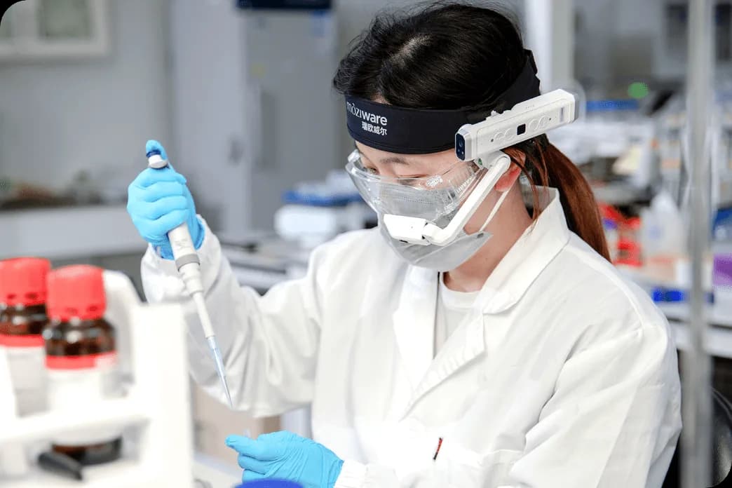 Moziware cımō worn by a woman in a lab, white module on a headband with the display arm positioned in front of her safety glasses.