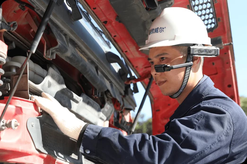 Moziware cımō worn under a hard hat by a man in coveralls, silver module beside his eye, working on a red truck engine.