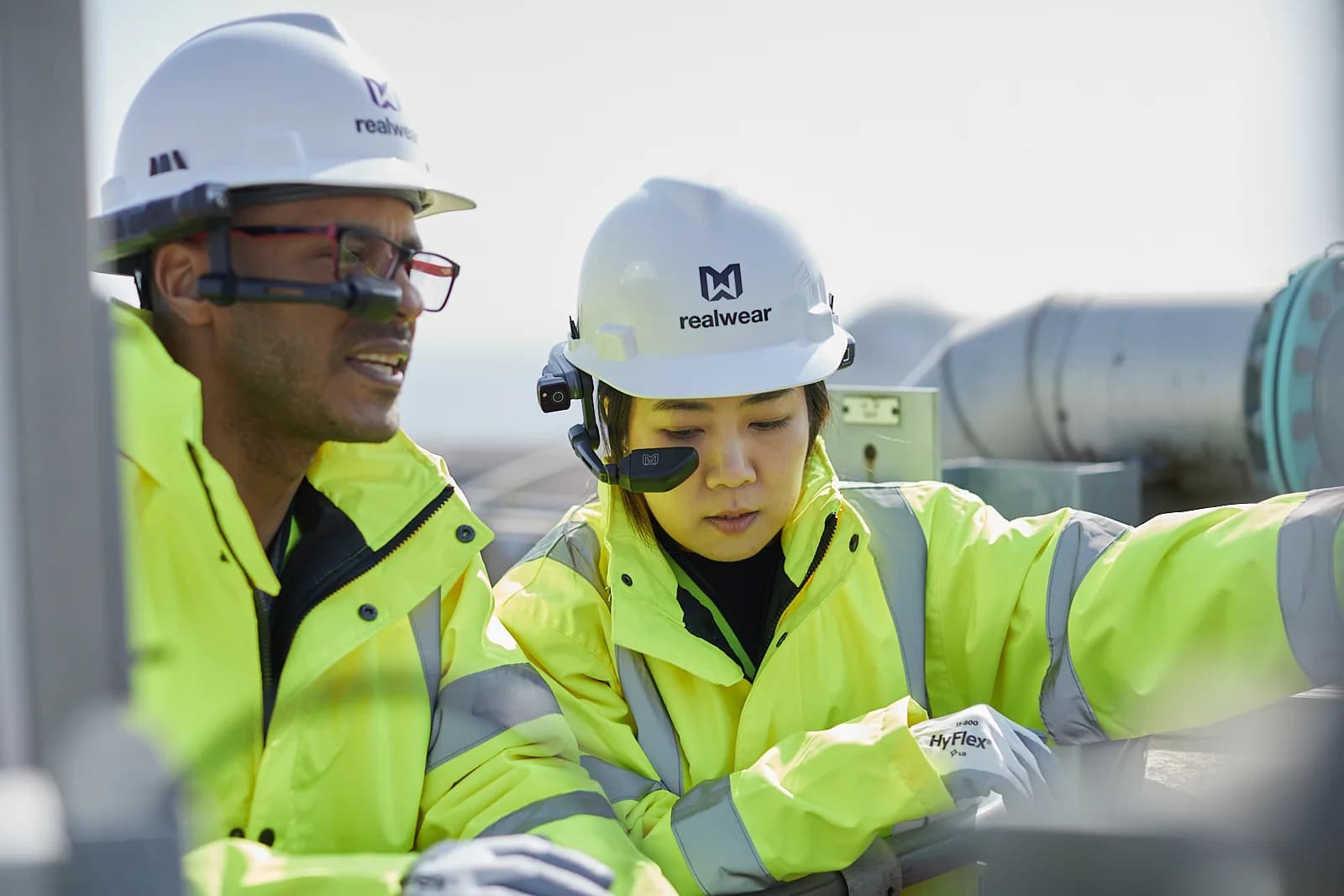 RealWear Navigator Z1 headset in red and black, worn over glasses by a worker in a yellow jacket and white hard hat outdoors.