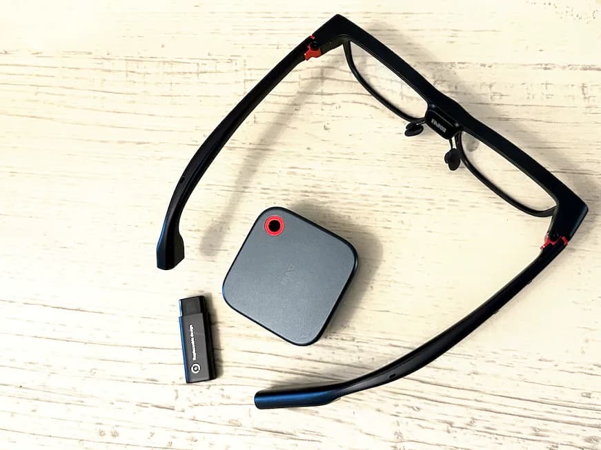XanderGlasses opened flat on a table beside a square gray battery pack and a small black remote.